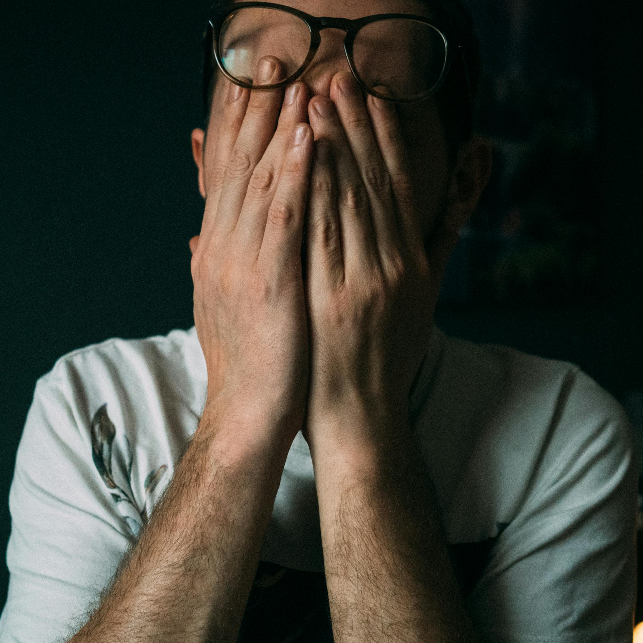Close-up of a man indoors with hands covering his face, expressing emotions like sadness or disappointment.